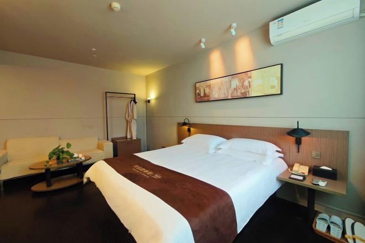 Habitación de hotel con cama, mesita de noche con lámpara, perchero, una silla y un cuadro en la pared.