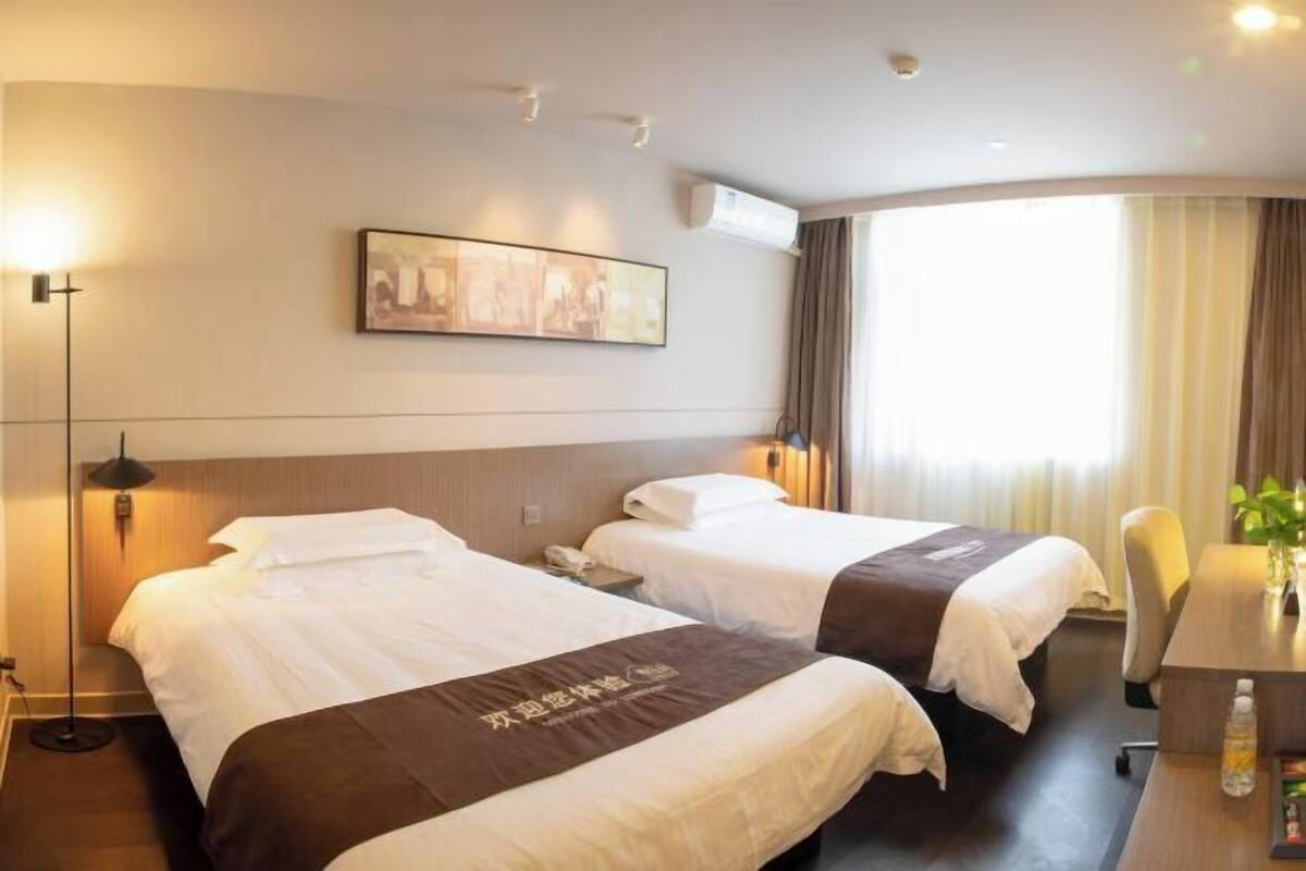 A hotel room with two beds, a desk, a chair, a lamp, and a painting on the wall.