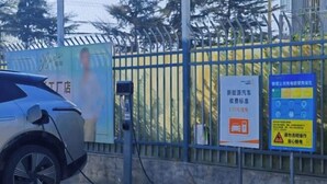 Une voiture en cours de recharge à une borne, avec une clôture et un panneau indicateur en arrière-plan.