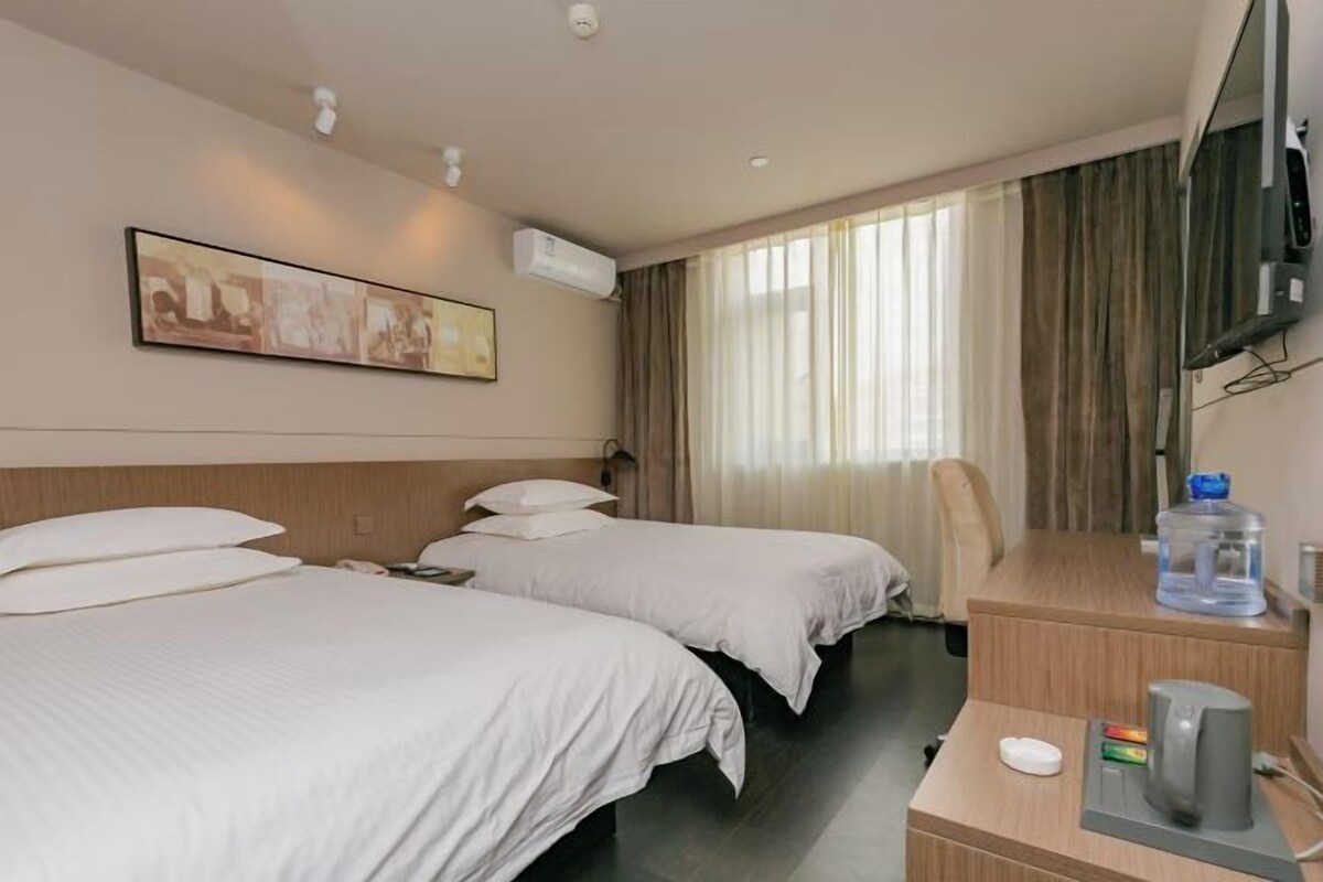 A hotel room with two beds, a desk with a kettle, a television, and an air conditioning unit.