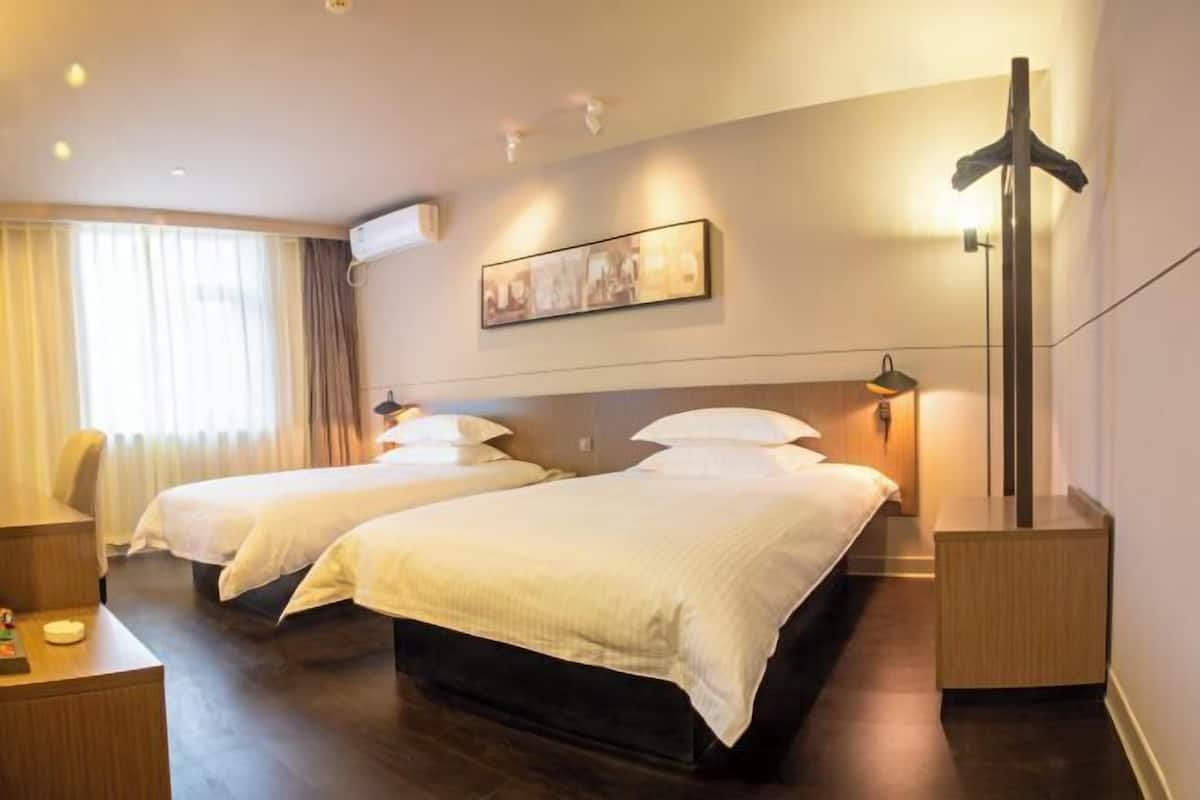 A hotel room with two beds, a desk, a chair, a coat rack, and a painting on the wall.