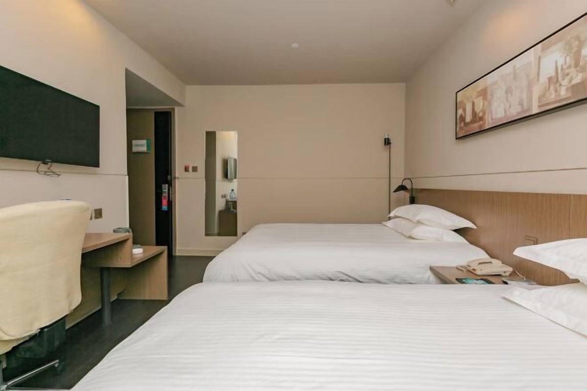Quarto de hotel com duas camas, uma escrivaninha, uma cadeira, uma televisão e um quadro na parede.