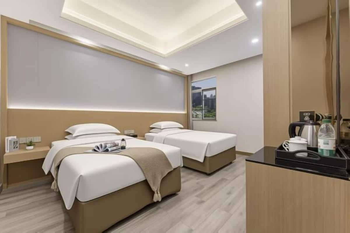 A modern hotel room with two beds, a bedside table, a mirror, and a small kitchenette.