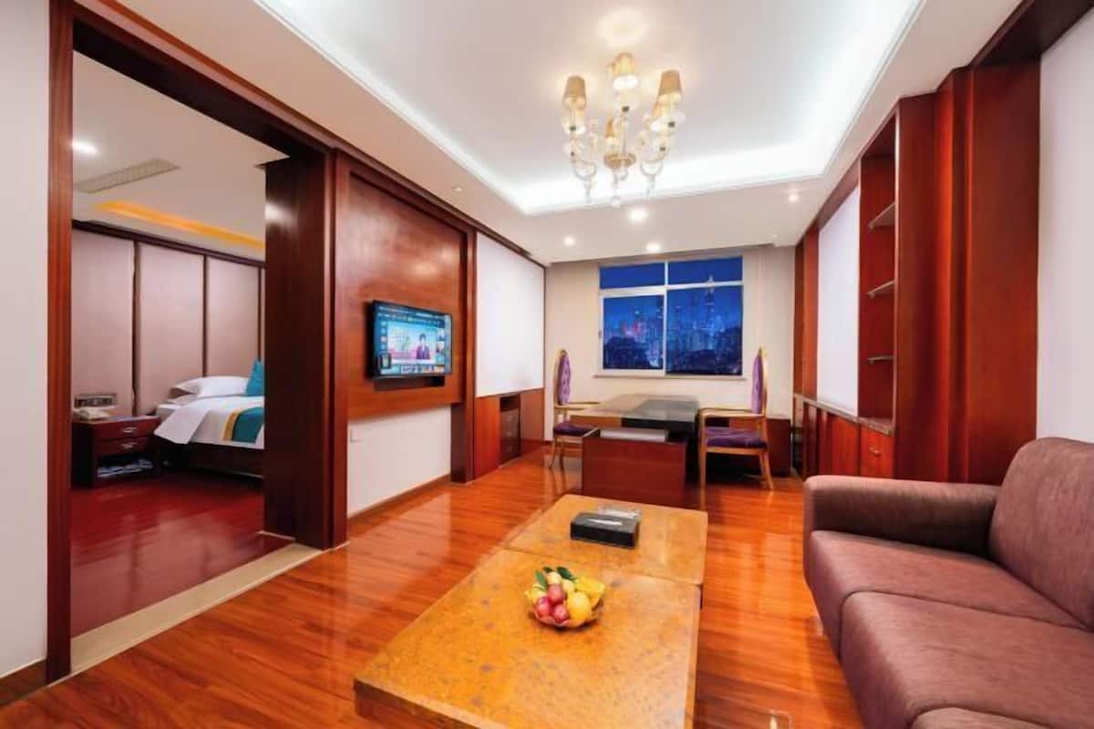 A hotel room with a wooden floor, a sofa, a TV, a desk with a chair, and a window with a city view.