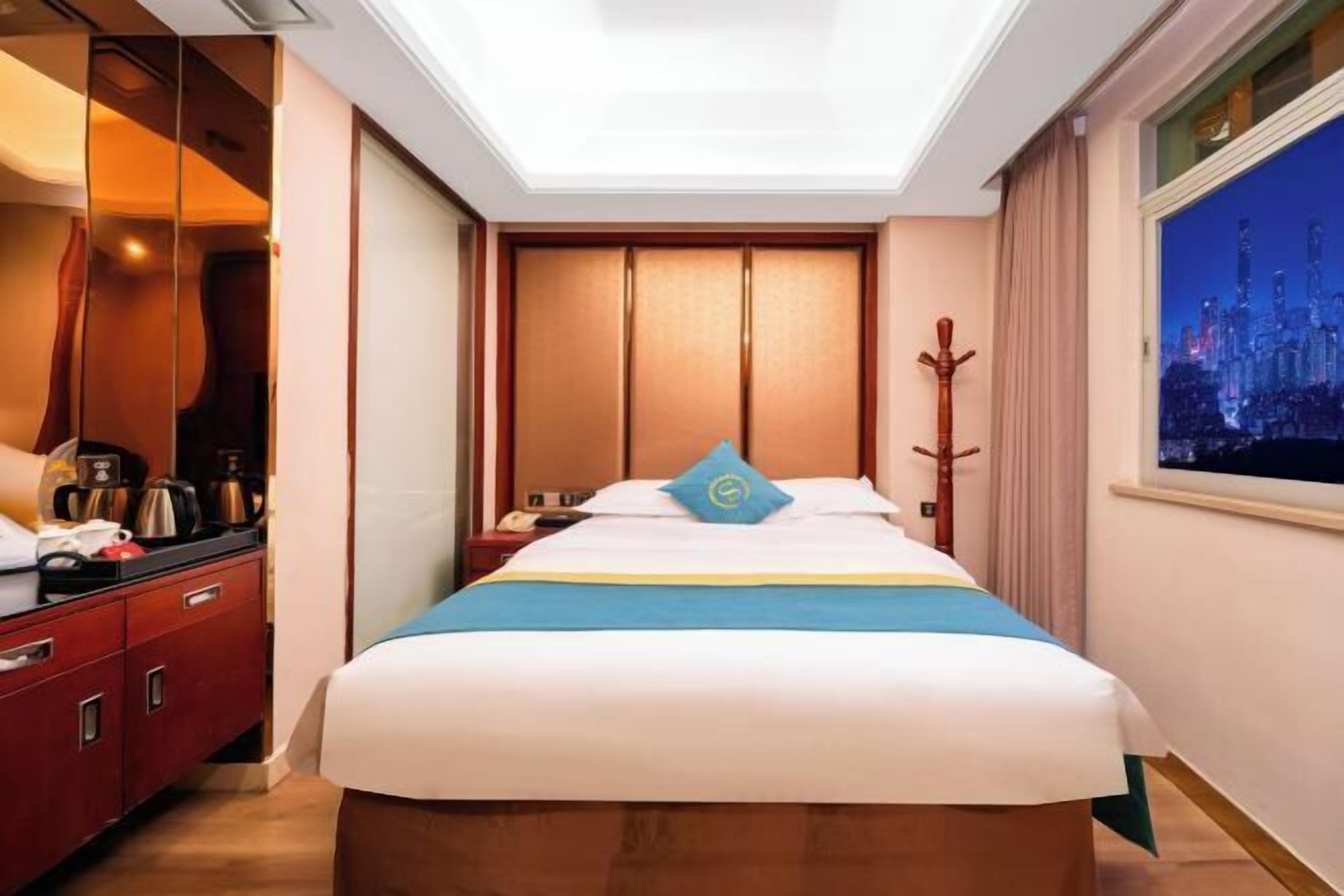A hotel room with a bed, a nightstand, a mirror, a wardrobe, and a window with a cityscape view.