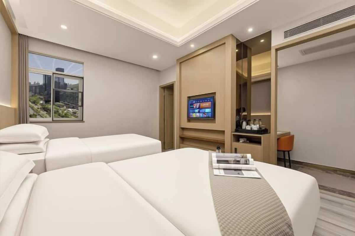 A modern hotel room with a large bed, a TV mounted on a wooden panel, a desk with a chair, and a window with a city view.