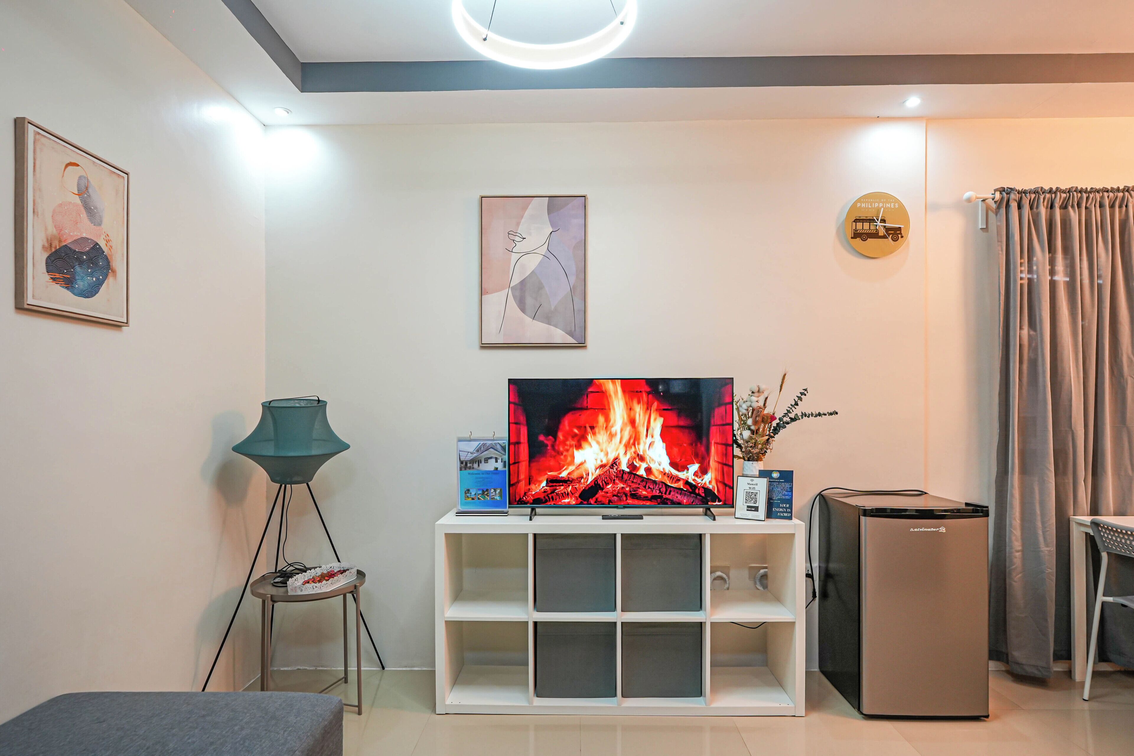 A modern living room with a flat-screen TV, a small fridge, a lamp, and artwork on the walls.