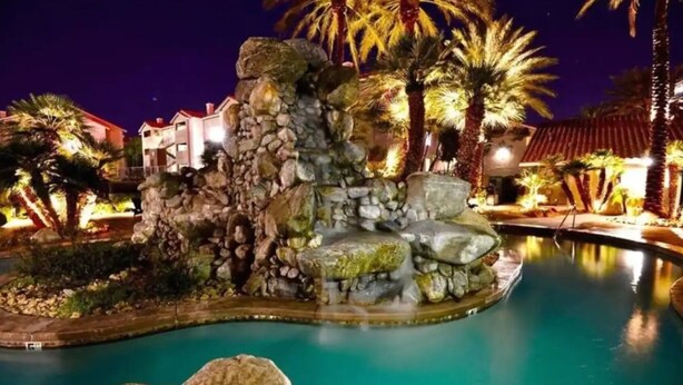 A nighttime view of a resort pool area with a rock formation centerpiece, palm trees, and illuminated buildings. - ★Strip views w/amenities! Pool, fitness center, W/D in-unit & FREE parking!★ (Las Vegas)