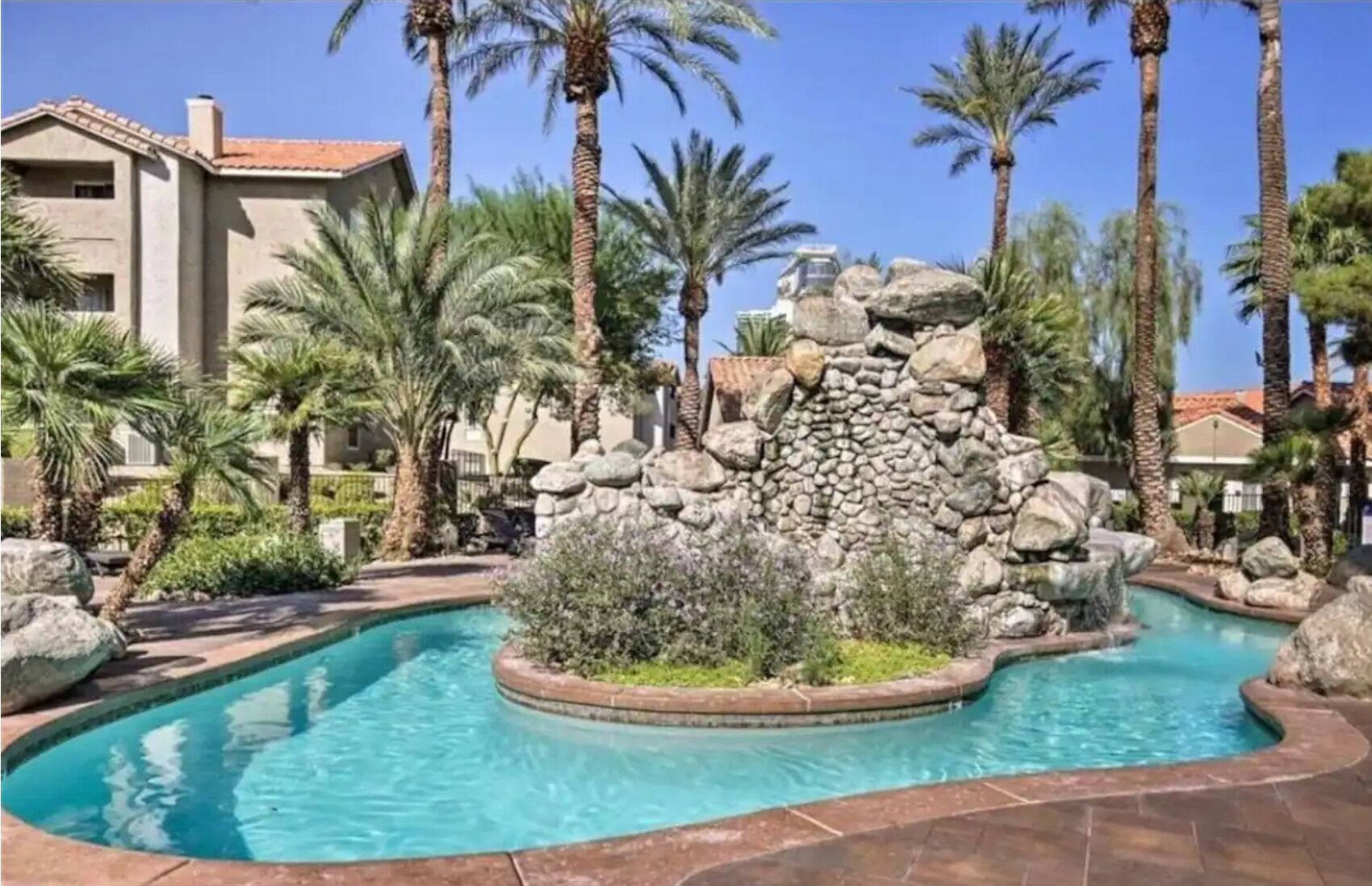 A pool area with a stone waterfall feature, surrounded by palm trees and a stone wall.