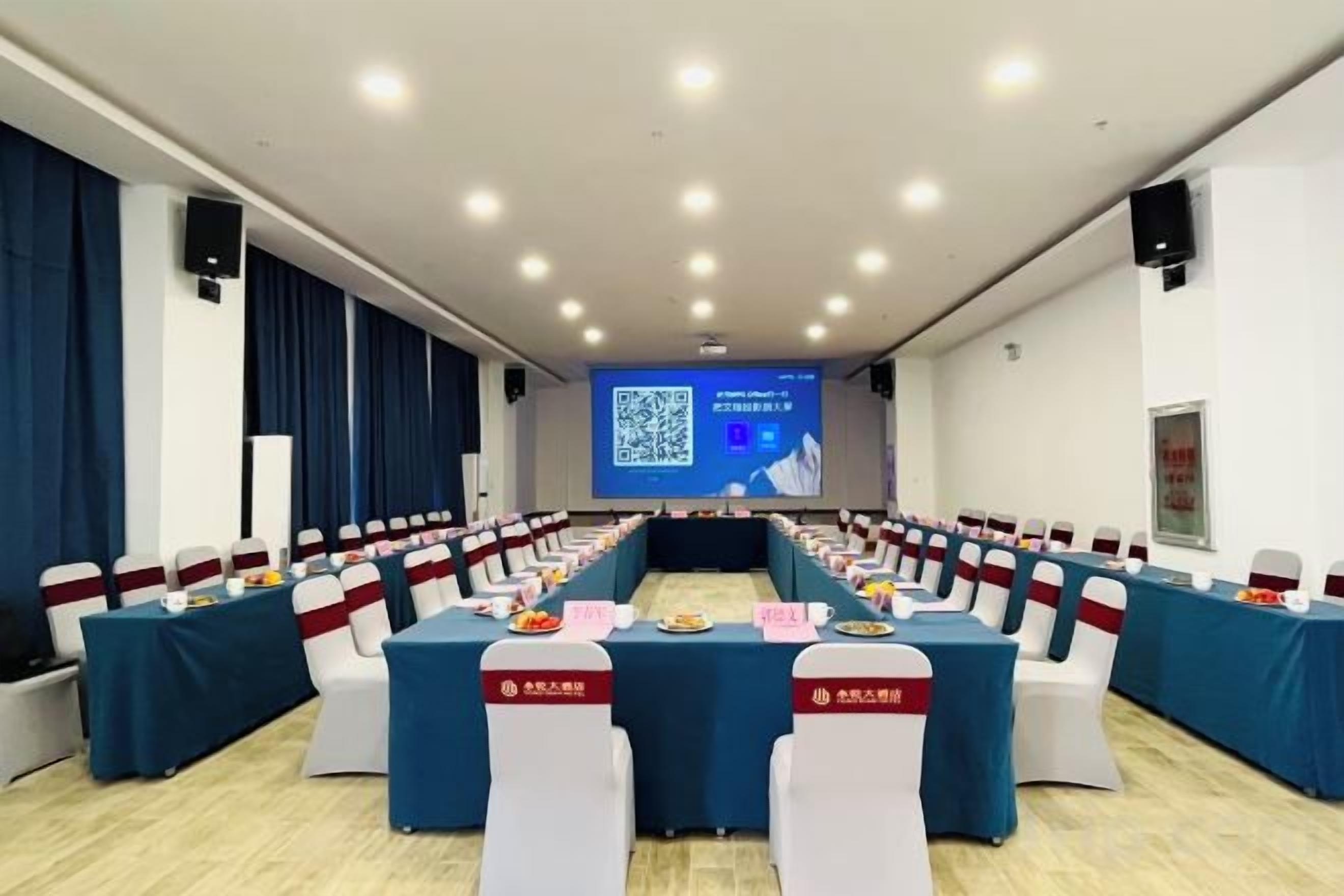 A conference room with tables set for a meeting, a screen at the front, and a speaker on the wall.