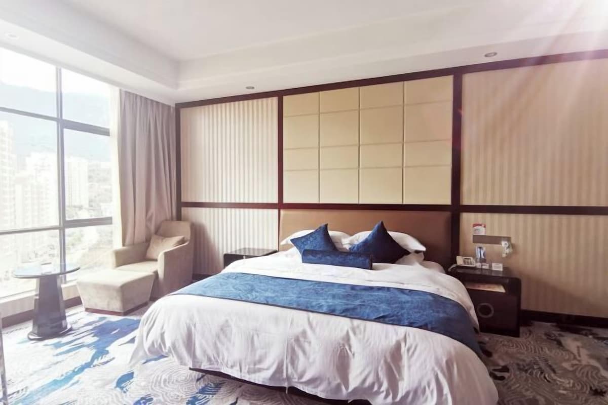 A hotel room with a large bed, a chair, a bedside table, and a view of the city through large windows.