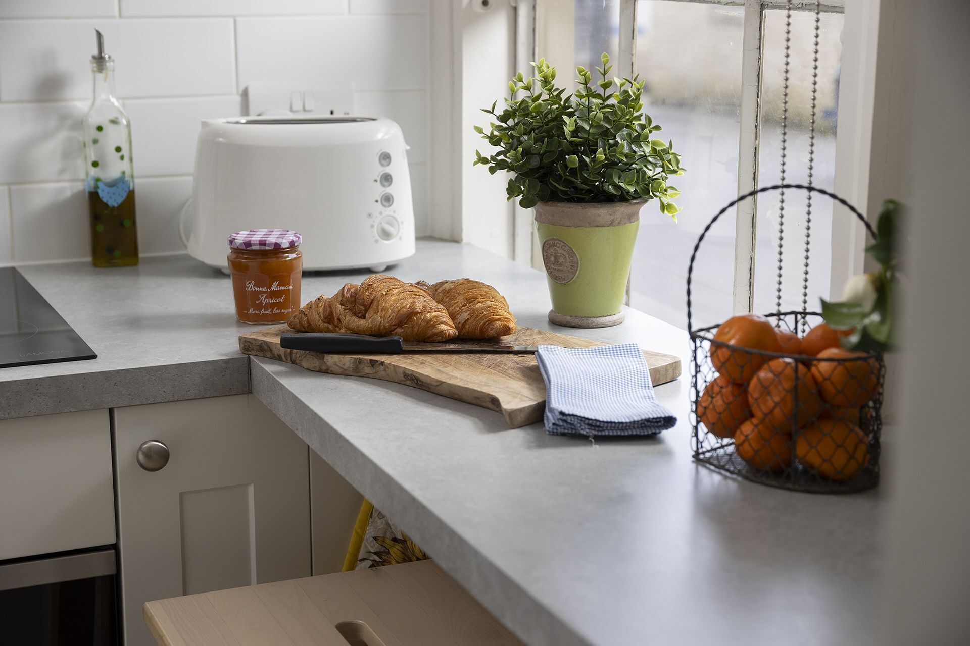 A kitchen counter with a toaster, a jar of honey, a plant, a basket of oranges, and a cutting board with a knife.