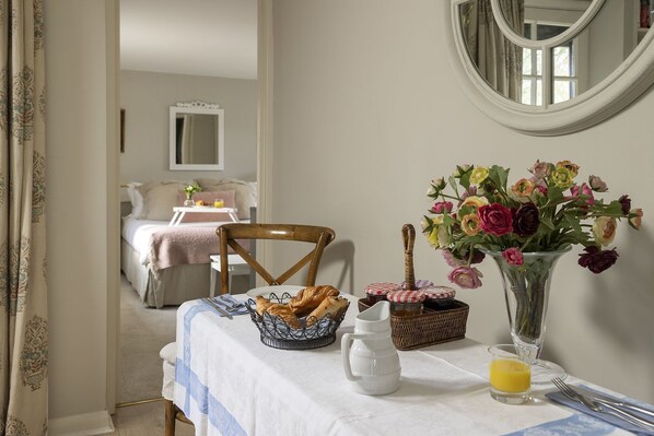 A breakfast setup with a table, a chair, a vase of flowers, a basket of bread, a pitcher, and a glass of orange juice.