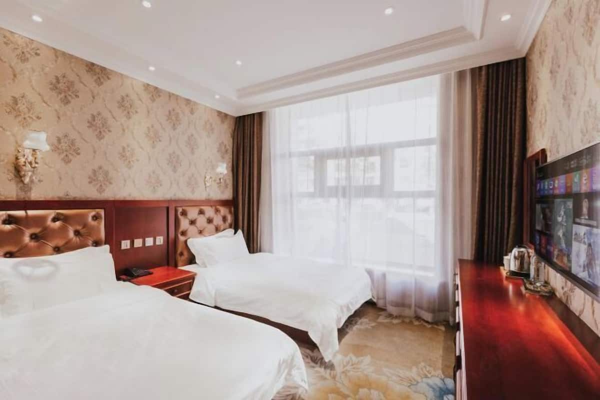 Ein Hotelzimmer mit zwei Betten, einem Holzkopfteil, einem großen Fenster mit Vorhängen, einem Wandfernseher und einer Holzkommode.