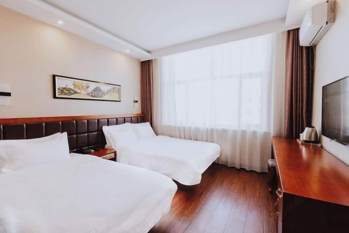 Ein Hotelzimmer mit zwei Betten, einem Holzkopfstück, einem Flachbildfernseher, einem Bild an der Wand und einem großen Fenster mit Vorhängen.