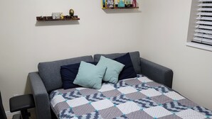 A compact bedroom with a sofa bed, a small desk, and a window with blinds.