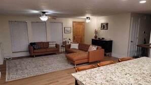A spacious living area with a wooden floor, a brown sofa set, a dining table with chairs, and a kitchen area with a granite countertop.