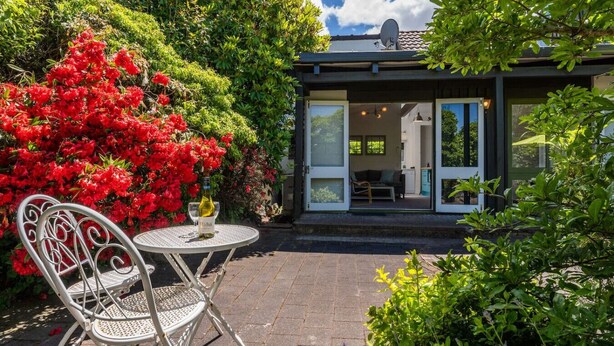 A modern house with a glass door leading to a living area, surrounded by lush greenery and a patio with a table and chairs. - Elegant Town House with Wi-Fi, central location, easy walk to town (Taupō)