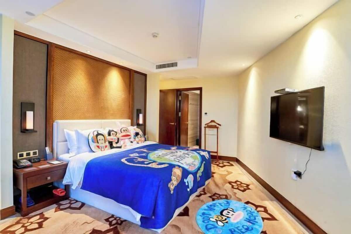 Una habitación de hotel con una cama, un televisor montado en la pared, una mesita de noche y una pequeña mesa redonda con un diseño de personaje de dibujos animados.