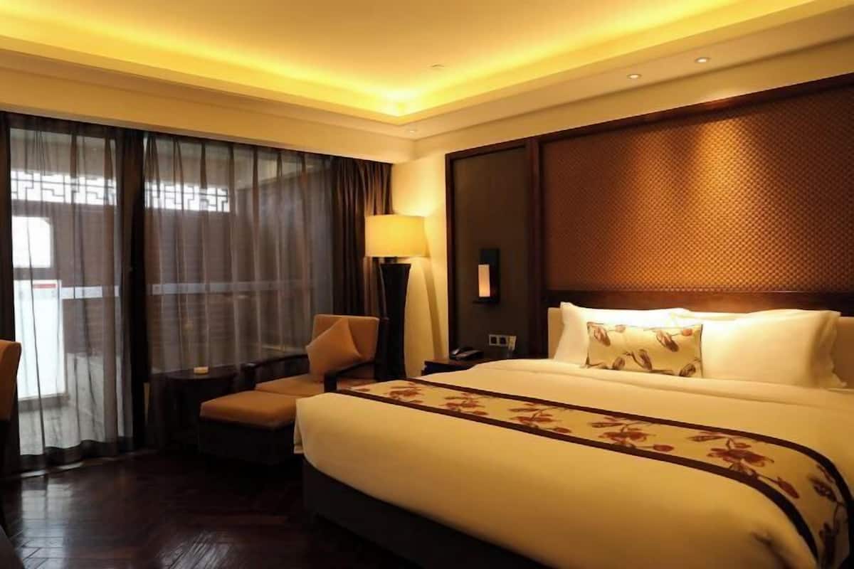 Habitación de hotel con una cama grande, una silla, una lámpara y vistas al exterior a través de cortinas translúcidas.