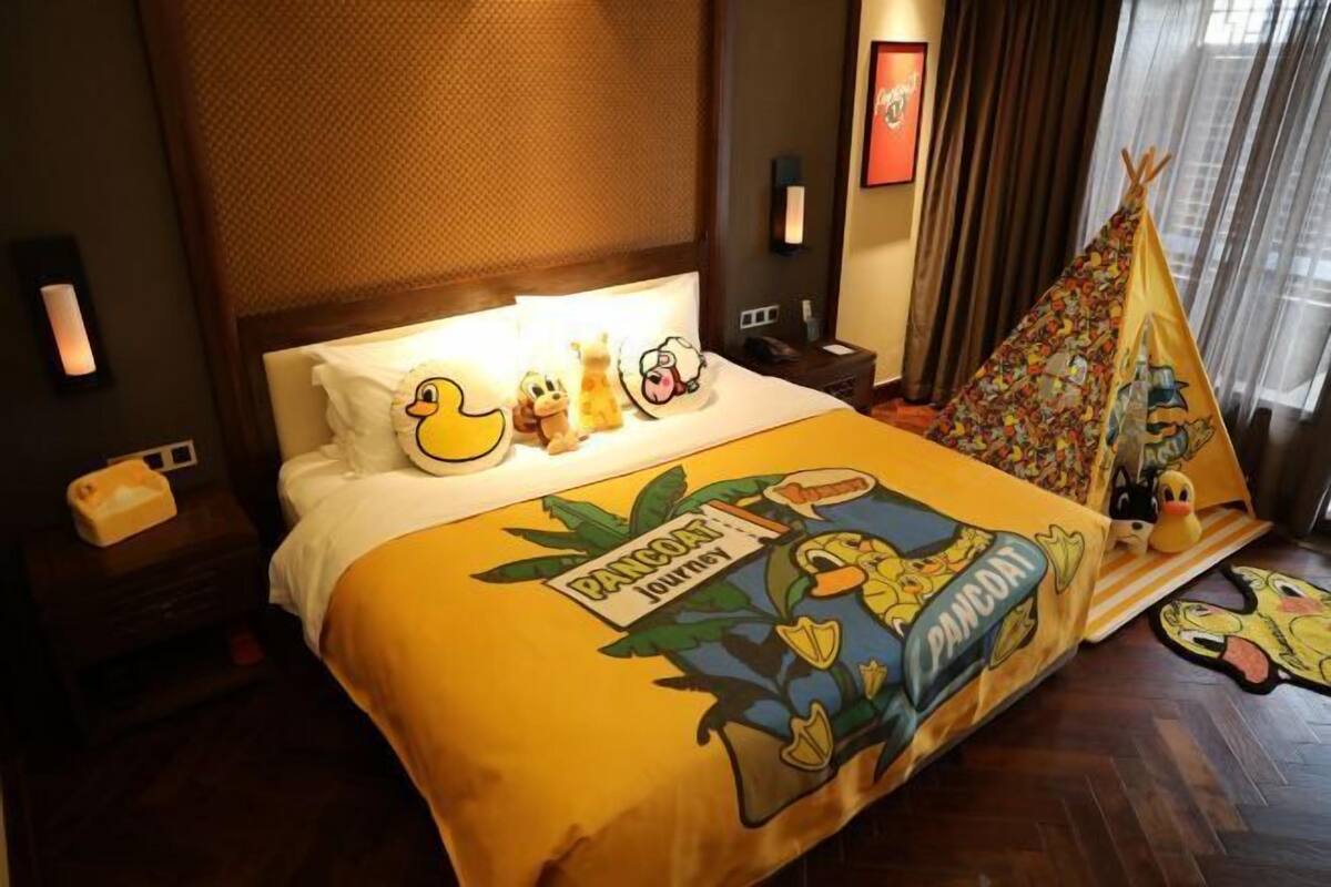 Habitación de hotel con una cama que tiene un edredón con temática de 'Pancake Journey', una mesita de noche con lámpara, y una ventana con cortinas.