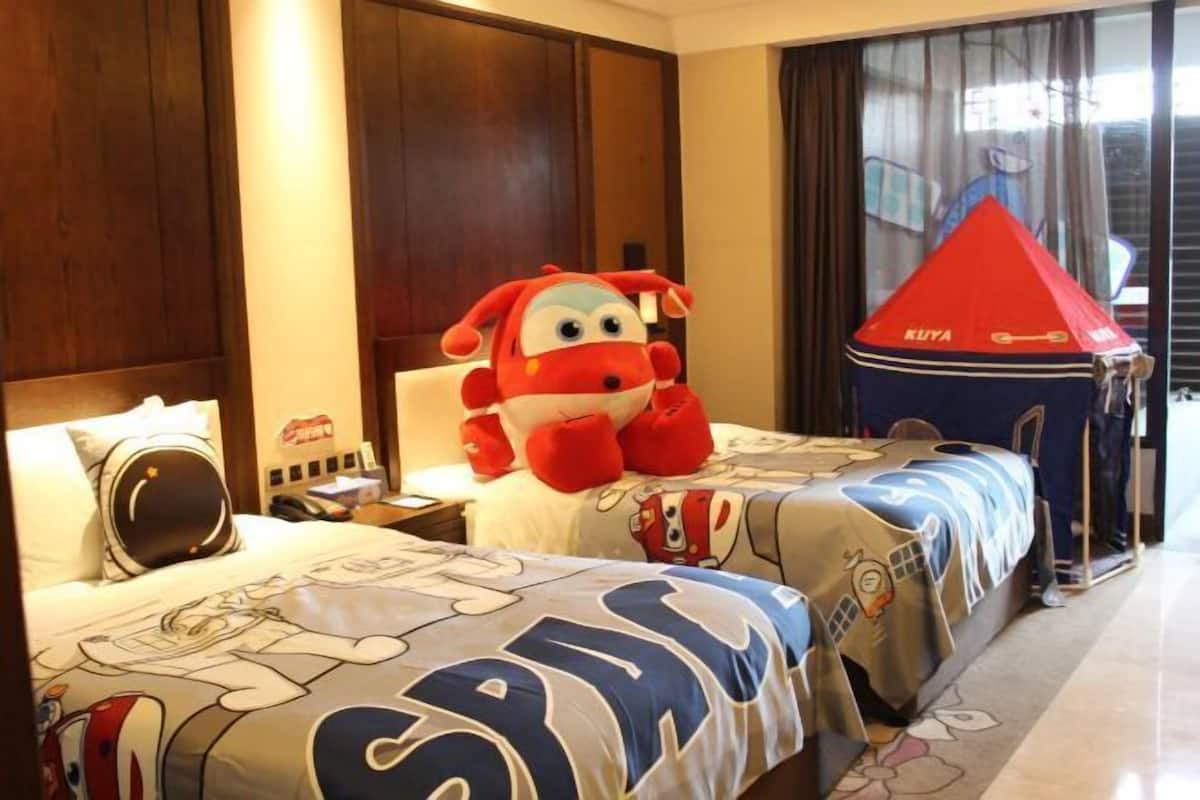 Habitación de hotel con dos camas, cada una con una colcha con motivos de dibujos animados, un peluche grande en una cama y un adorno en forma de paraguas rojo en la otra.