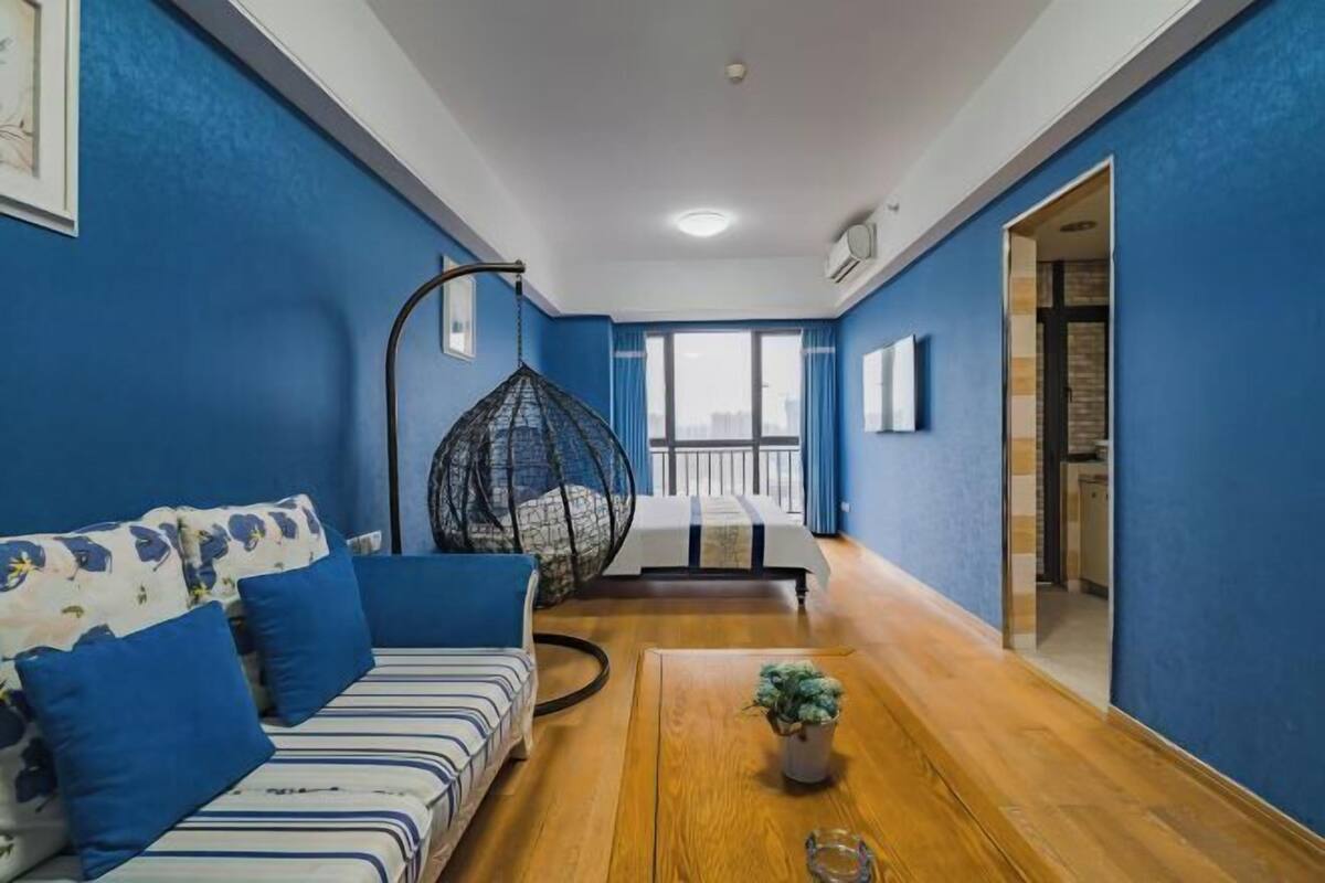 Ein Zimmer mit blauer Wand, Holzboden, einem gestreiften Sofa mit blauen Kissen, einem Hängekorbstuhl und einem Bett mit Blick ins Freie.