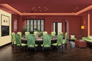 A dining area with a round table, green upholstered chairs, a large window with patterned curtains, and a red accent wall.