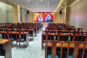 Un salón de conferencias con filas de mesas y sillas de madera, una pantalla grande al frente y botellas de agua sobre las mesas.