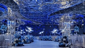 Un espacio para eventos formales con un escenario central, arreglos florales blancos y un techo con superficies reflectantes.