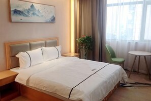 A hotel room with a bed, a nightstand, a chair, a small table, and a window with curtains. - Hefu International Hotel (Nanzhang)