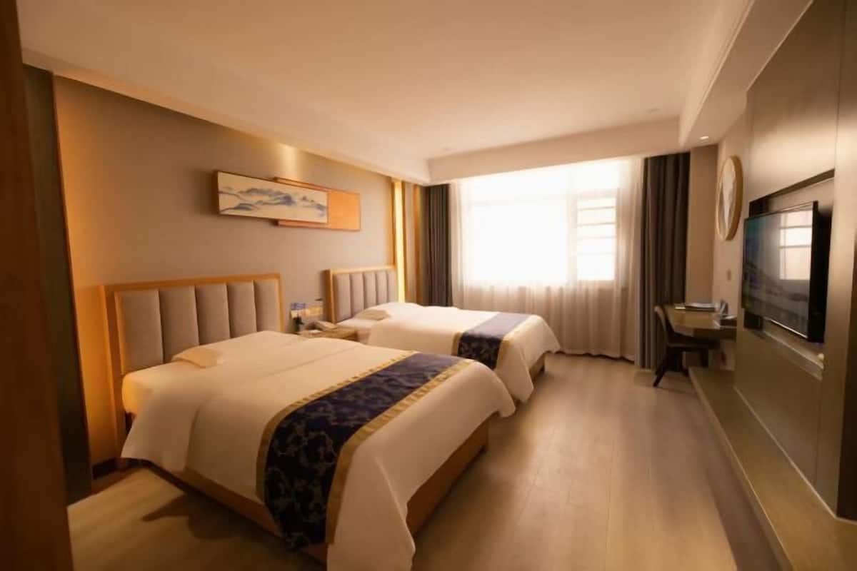 Ein Hotelzimmer mit zwei Betten, einem Schreibtisch, einem Fernseher und einem großen Fenster mit Vorhängen.