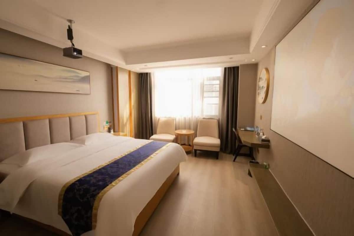 Ein Hotelzimmer mit einem großen Bett, zwei Sesseln, einem kleinen Tisch, einem Projektor und einem großen Fenster mit Vorhängen.