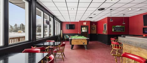 Una sala de billar con mesa de pool, sillas rojas y zona de bar.