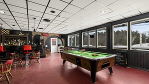Une table de billard dans une pièce au sol rouge, un coin bar avec une enseigne Coca-Cola et de grandes fenêtres donnant sur des arbres.