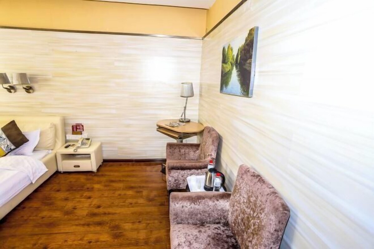 A hotel room with a bed, a nightstand with a phone, a small table with a lamp, and two armchairs.