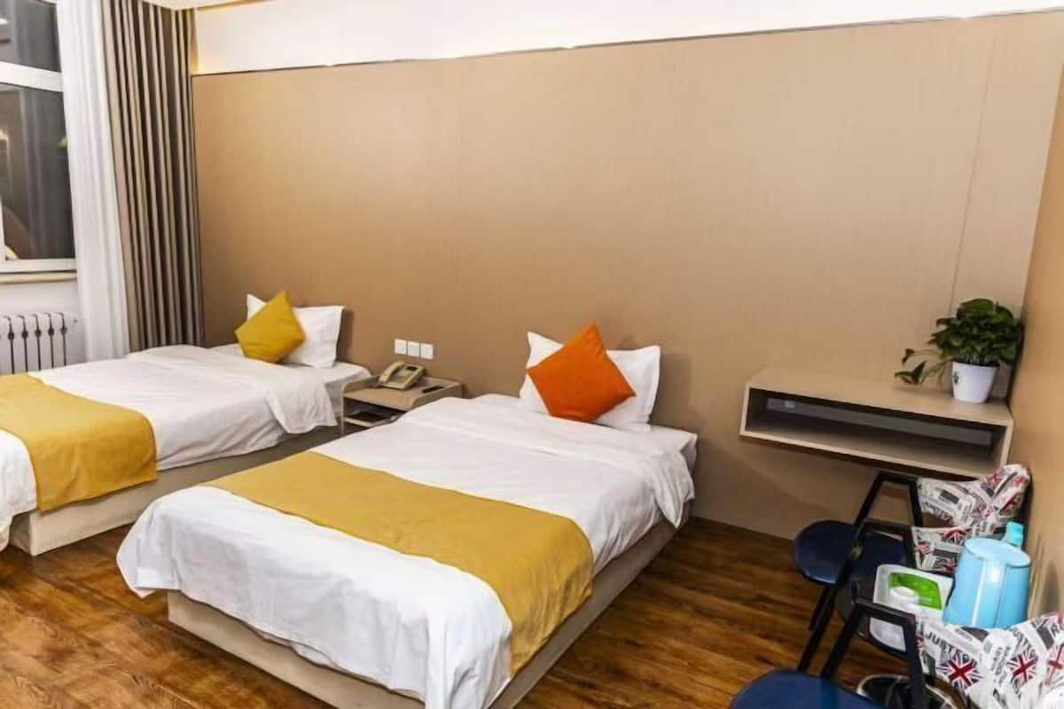 Hotellihuone, jossa on kaksi sänkyä. Kummassakin sängyssä on valkoiset vuodevaatteet ja päällä keltainen torkkupeitto. Sängyn vieressä on yöpöytä, jossa on puhelin, ja sen yläpuolella on pieni hylly.