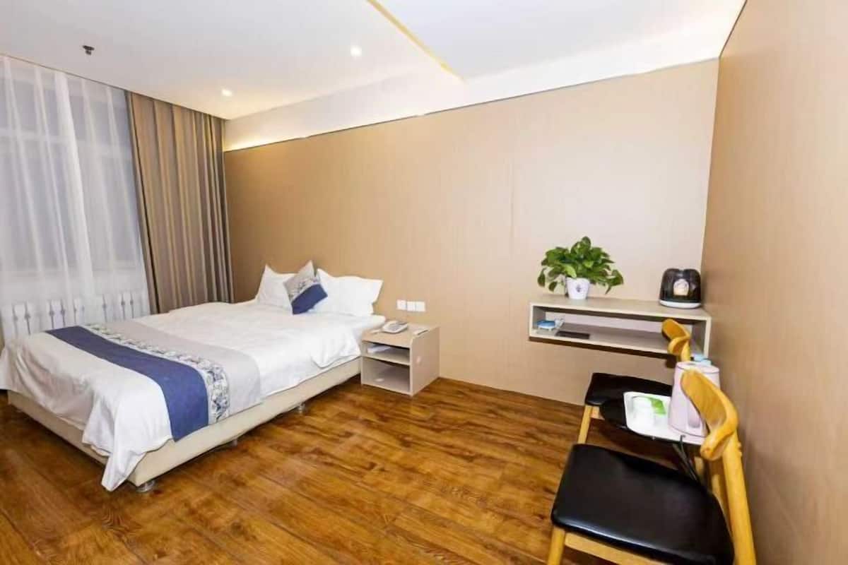 Hotellihuone, jossa on sänky, työpöytä, tuoli, pieni hylly, jossa on kasvi, ja ikkuna, jossa on verhot.