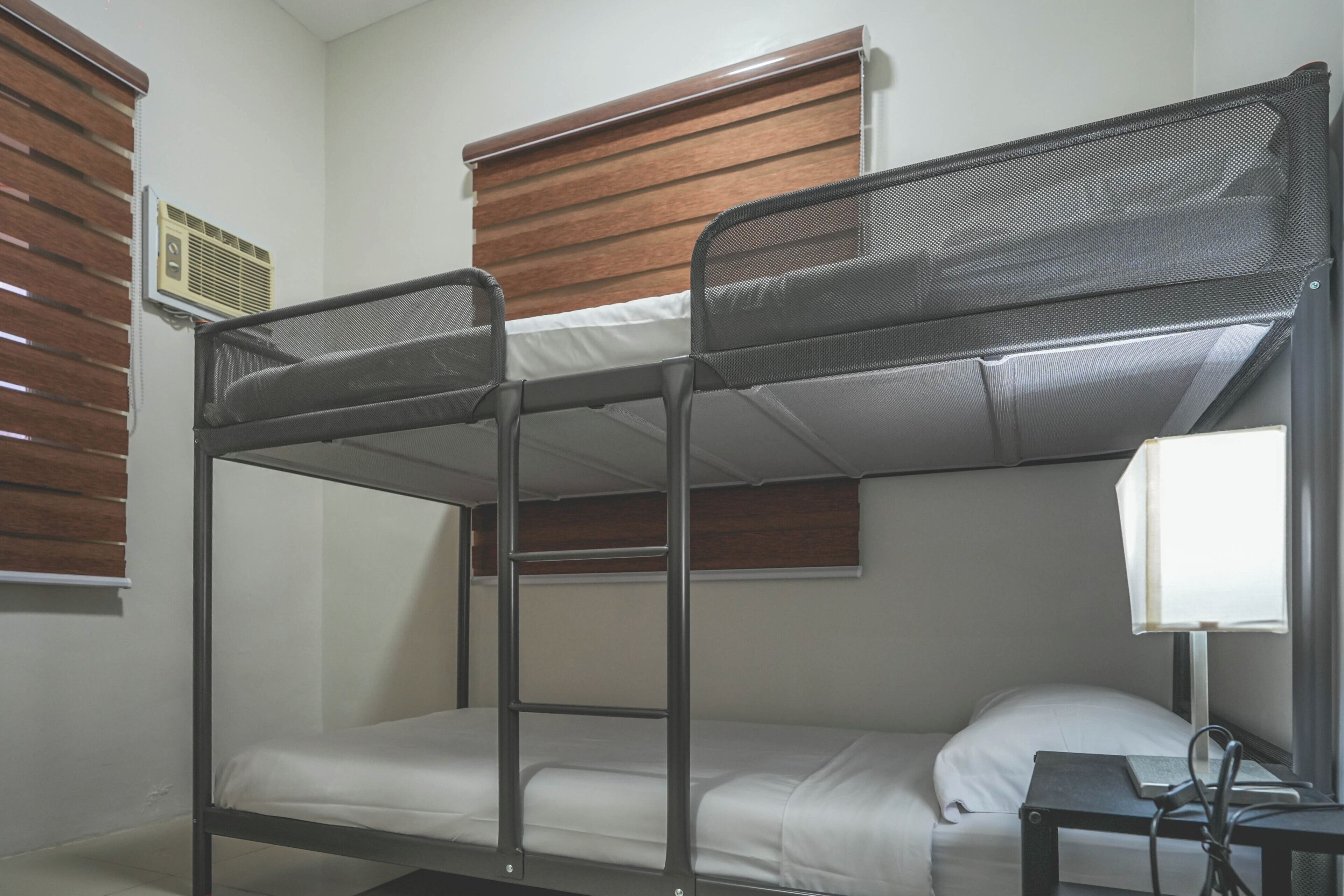 A bunk bed room with a desk and lamp, wooden blinds, and a wall-mounted air vent.