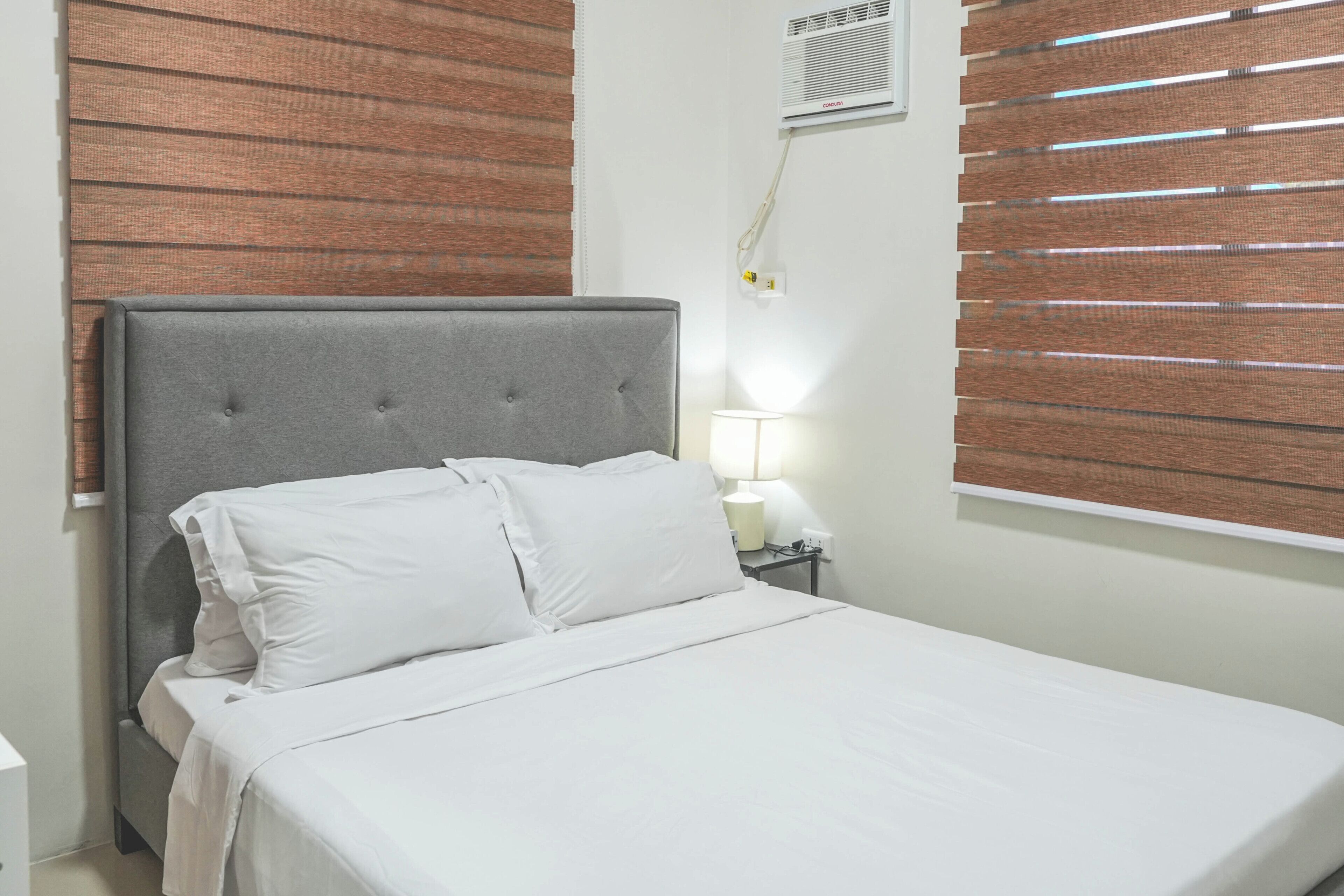 A bed with a grey upholstered headboard, white bedding, and a bedside lamp.
