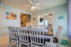 A dining area with a wooden table and white chairs, a ceiling fan, and a painting of a sunset. - Lo-Key 1: Discover Your Slice of Paradise, Charming Marathon Home, Pool, Deck, Patio & Dog-Friendly! (Marathon)