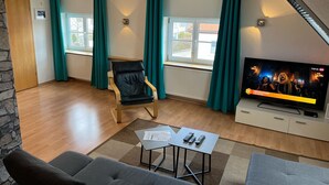 A modern living room with a sofa, a chair, a coffee table, and a TV. There are windows with curtains, a radiator, and a stone wall. - HoLiDay HouSE Allgäu (Ottobeuren)