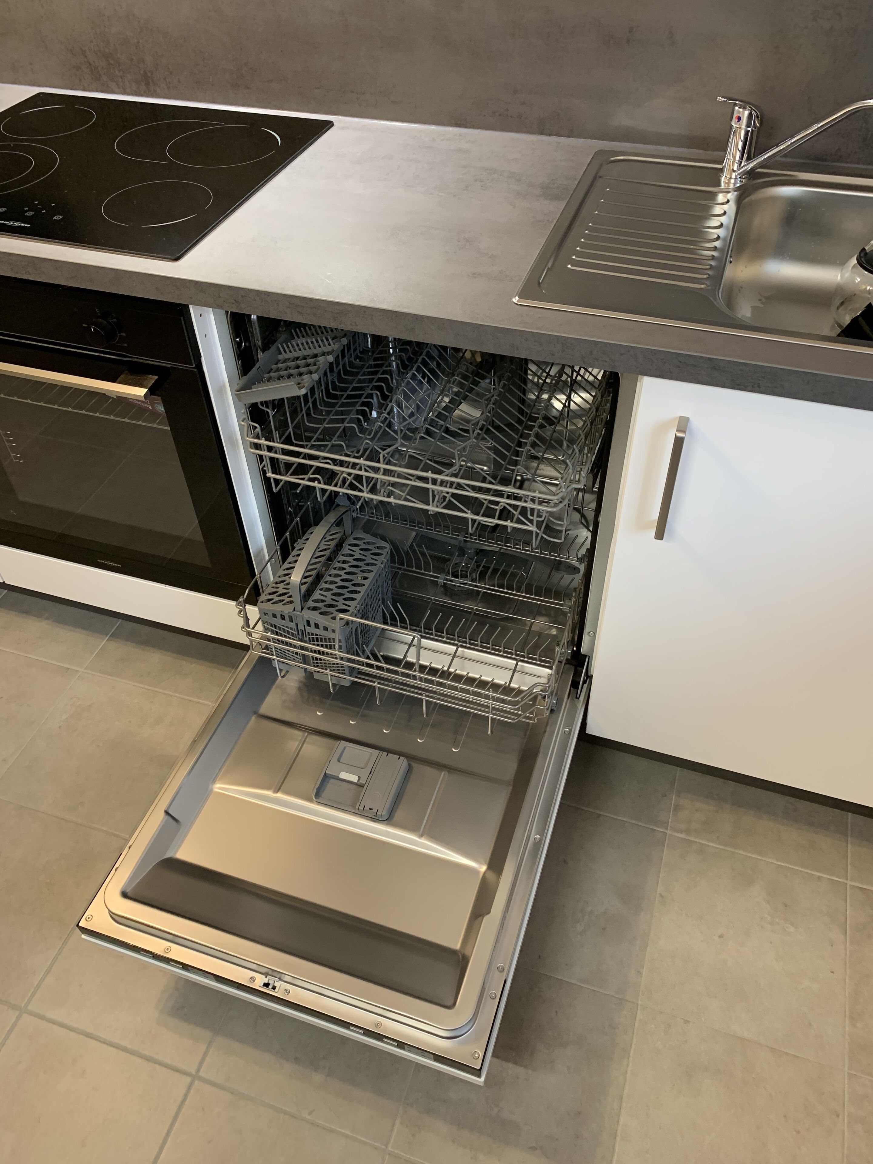 An open dishwasher in a kitchen with a built-in oven and a stainless steel sink.