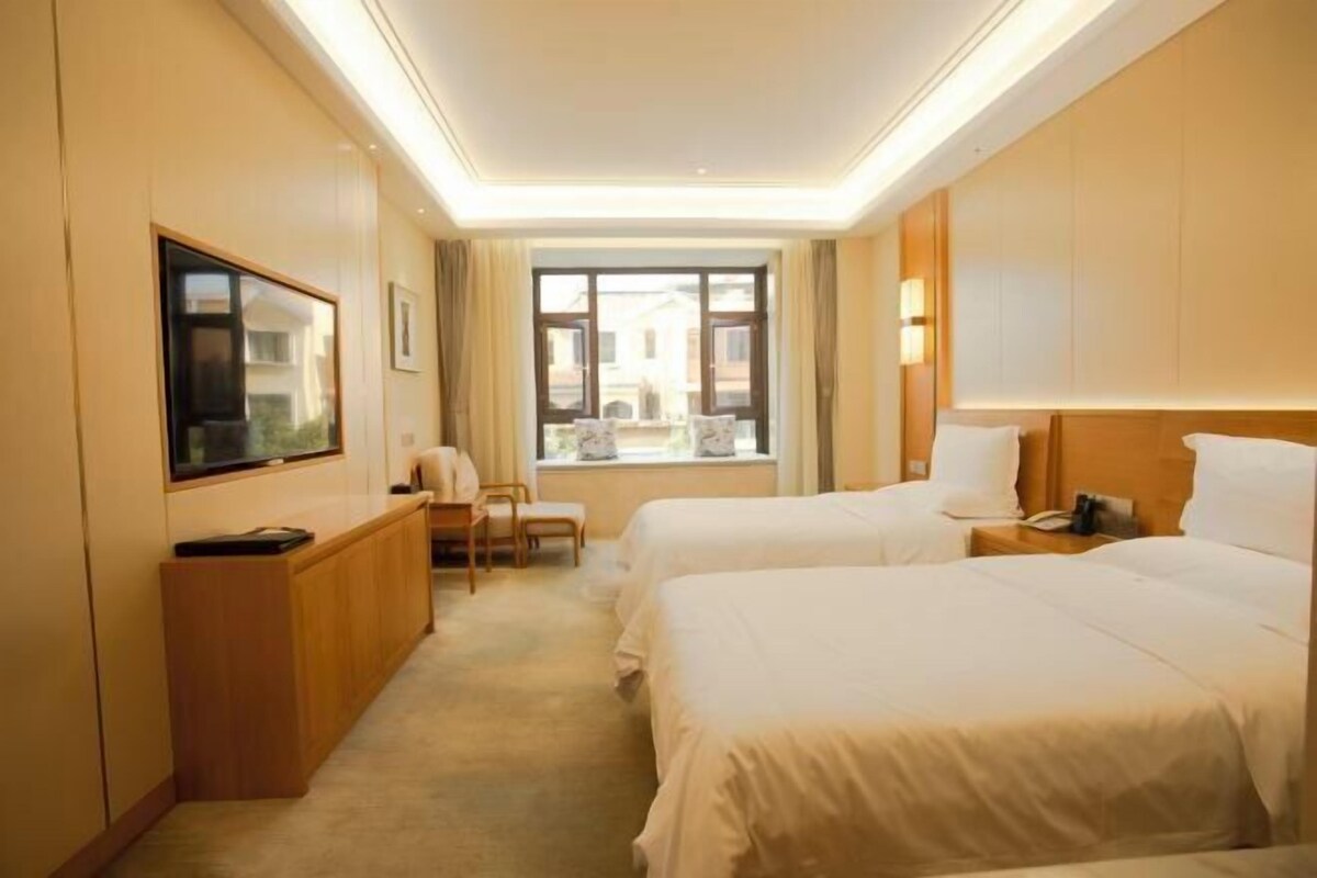 Ein Hotelzimmer mit zwei Betten, einem Schreibtisch, einem Stuhl und einem großen Fenster mit Blick auf Gebäude.