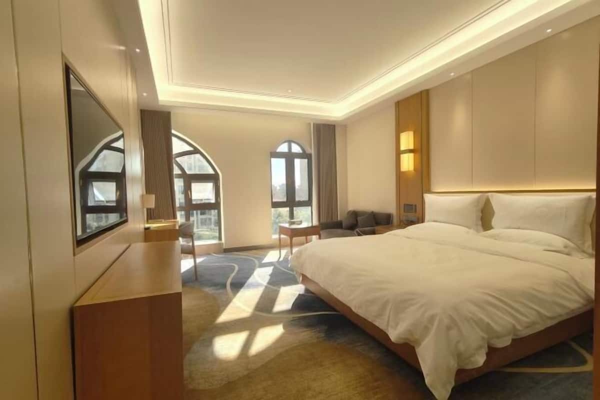 Ein modernes Hotelzimmer mit einem großen Bett, einem Schreibtisch und einem Fernseher. Zwei Fenster bieten Blick auf die Stadt.
