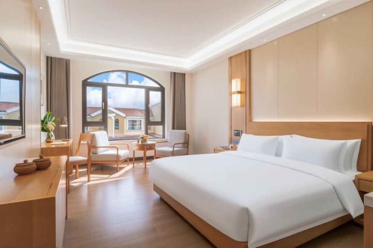 Une chambre d’hôtel avec un grand lit, un bureau, deux chaises et une vue sur les immeubles, visible à travers une grande fenêtre.