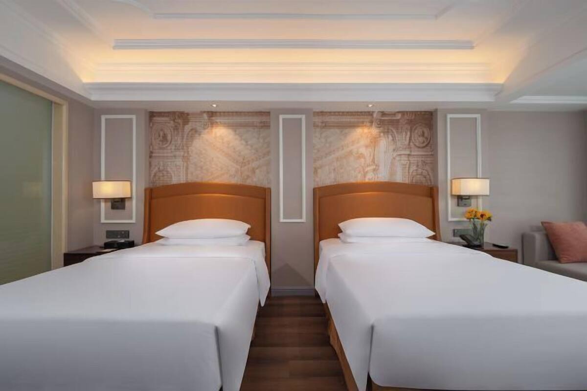 A hotel room with two single beds, a nightstand with a lamp, a small table with a vase of flowers, and a chair.
