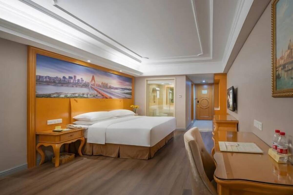 A hotel room with a large bed, a desk, a chair, a painting on the wall, and a door leading to another room.