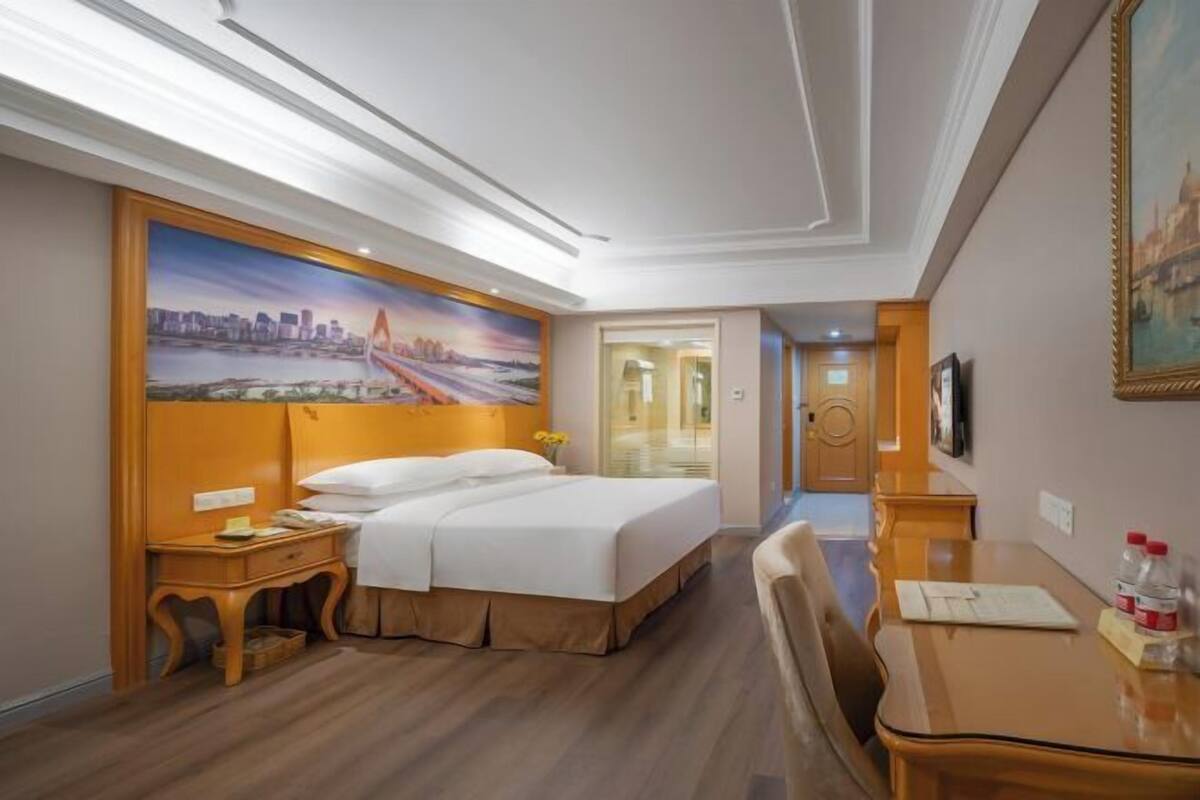 A hotel room with a bed, bedside table, a desk with a chair, a painting on the wall, and a door leading to another room.