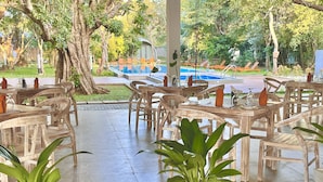 Uma área de refeições ao ar livre com mesas e cadeiras de madeira, cercada por plantas em vasos e vista para uma piscina.